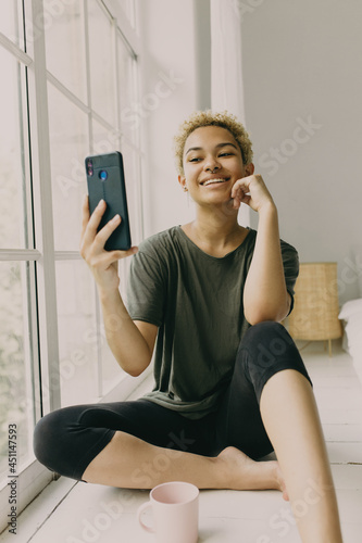 Picture of mixed race blonde girl, phone and pink coffee cup on floor near panoramic window. Curly-headed woman holding smartphone and posing for selfie, having toothy smile, touching cheek with hand