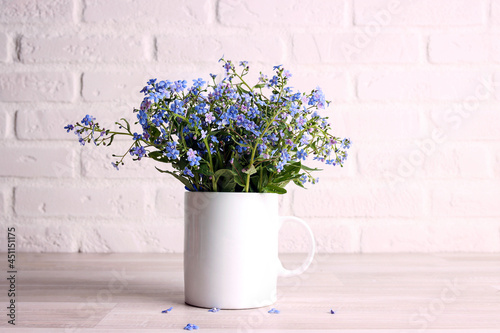 White coffee mug with bouquet of forget-me-not flowers on the white brick wall background.