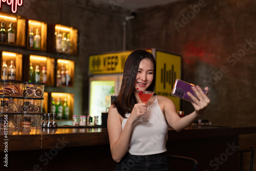 Nightlife concept a sexy girl with smiling face doing selfie on her smartphone while hold a vivid pink champaign in the night bar