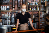 The waitress for the counter waiting to order beverages Portrait of a young beautiful waitress with protective face mask in a modern uniform standing with a smile in a cafe leaning against the counter