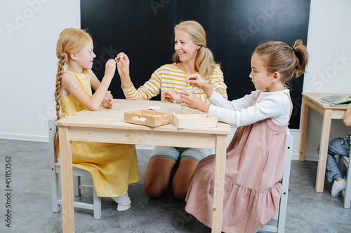 Smiling teacher helping girls to build 3d shapes from plasticine and toothpicks. They all sitting behind the same small square table. Girls wear nice long concervative dresses. photo