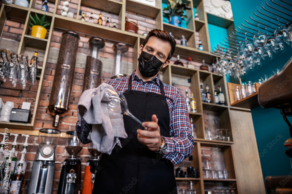 Cleaning the bar in the restaurant. An adult handsome man in a waiter's uniform stands behind the bar and wipes freshly washed wine glasses with a cloth. Restaurant work and corona virus