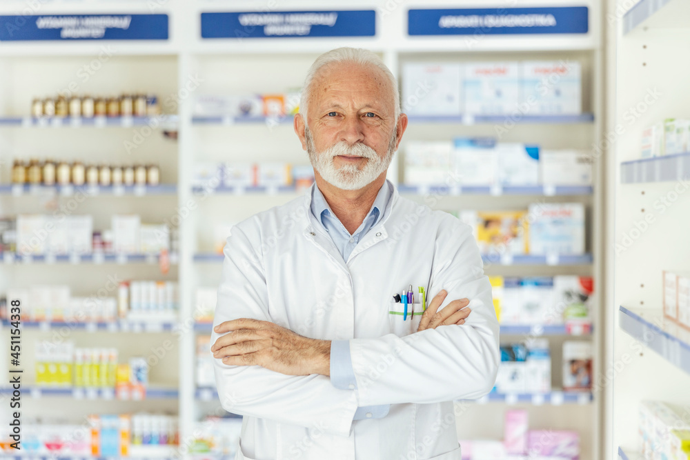 Portrait of a pharmacist in a pharmacy. Close-up shot of a male pharmacist in uniform and gray hair and beard crossed arms in the middle of the pharmacy in front of a shelf with medicines Proud worker