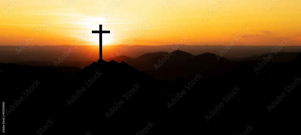 Religious grief landscape background banner panorama - View with black silhouette of mountains, hills, forest and cross / summit cross, in the evening during the sunset, with orange colored sky..