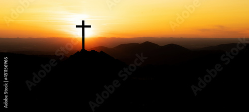 Fotografering Religious grief landscape background banner panorama - View with black silhouette of mountains, hills, forest and cross / summit cross, in the evening during the sunset, with orange colored sky