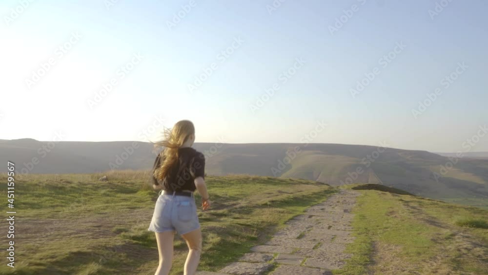 Stabilised shot of young blonde woman jogging along the path on top of Mam Tor, Castleton, Peak District, England with view of green rolling hills and blue skies. Stabilised shot of young blonde woman jogging along the path on top of Mam Tor, Castleton, Peak District, England with view of green rolling hills and blue skies.