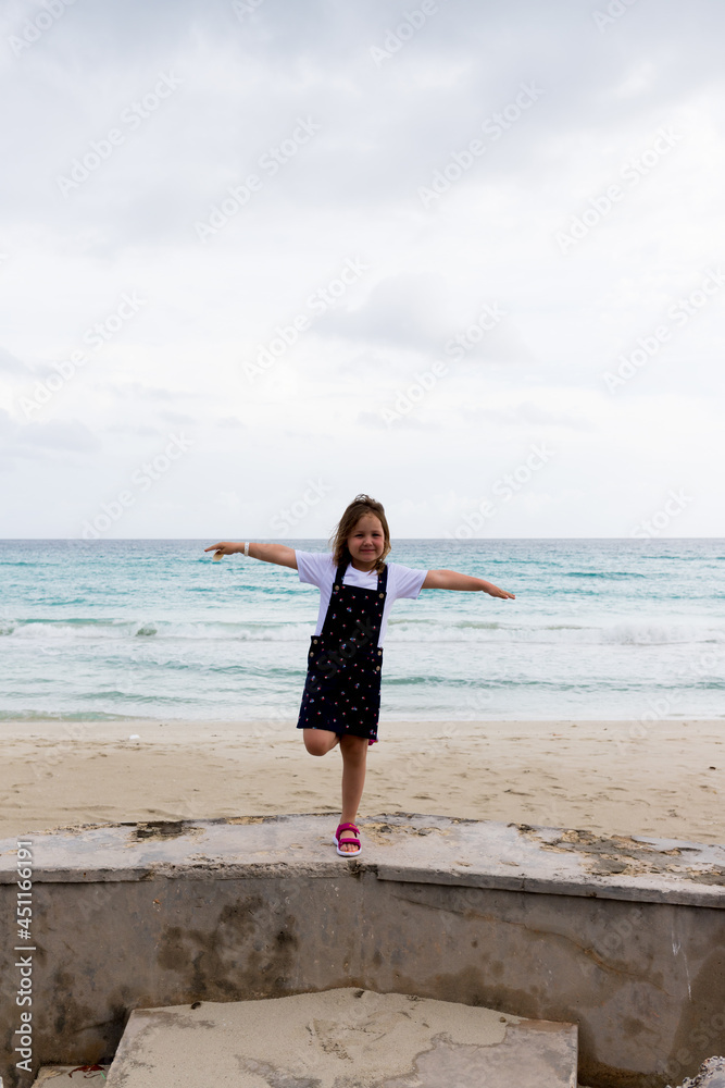 the child stands on one leg, arms outstretched to the sides against the background of the sea
