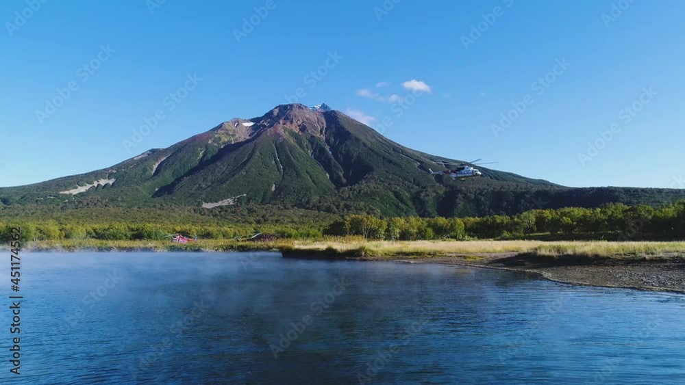 Flying over a lake in the haze in Kamchatka, Russia. A volcanic mountain on the background. On the right, the helicopter takes off and the camera gradually turns towards it.