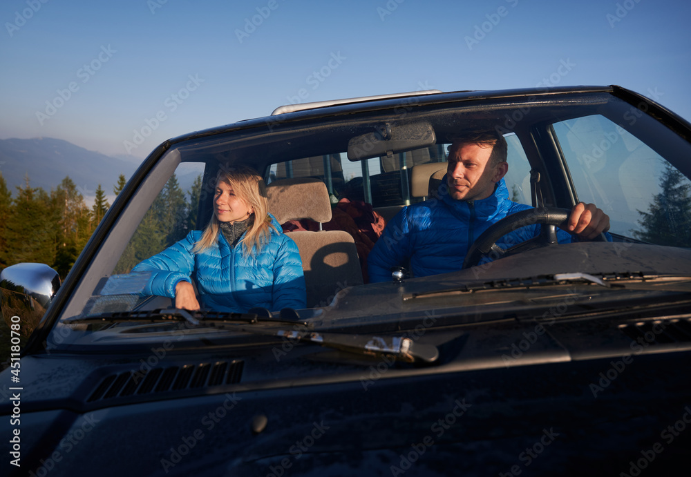Charming female traveler enjoying beauty of nature and smiling while man driver keeping hands on steering wheel and driving the car. Young couple travelling together in automobile in mountains. Charming female traveler enjoying beauty of nature and smiling while man driver keeping hands on steering wheel and driving the car. Young couple travelling together in automobile in mountains.
