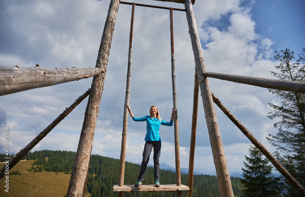Slender blonde woman standing on seat of very large wooden frame swing, holding on to side supports and smiling happily. On the background blue sky with gray clouds, forest and mountain hills. Slender blonde woman standing on seat of very large wooden frame swing, holding on to side supports and smiling happily. On the background blue sky with gray clouds, forest and mountain hills.