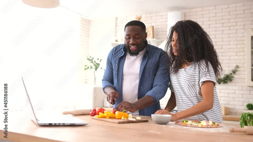 Joyful mixed race couple enjoying cooking with a video tutorial, happy African boyfriend and biracial girlfriend preparing meal in front of the laptop, a guy is biting slice of tomato and smiles Joyful mixed race couple enjoying cooking with a video tutorial, happy African boyfriend and biracial girlfriend preparing meal in front of the laptop, a guy is biting slice of tomato and smiles