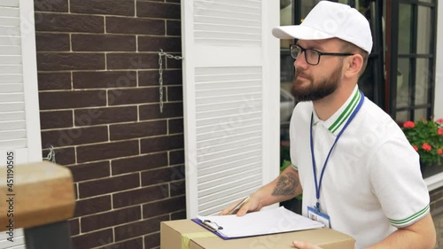 Young bearded postman in white cap and casual outfit carrying box, ringing at doorbell. Tattooed cheerful male courier comparing house number with data in folder. Delivery concept.