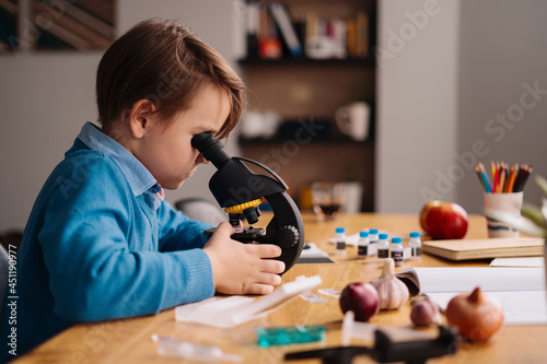 Distance online education, internet learning. First grade boy studying at home using microscope, making notes, biology online lesson