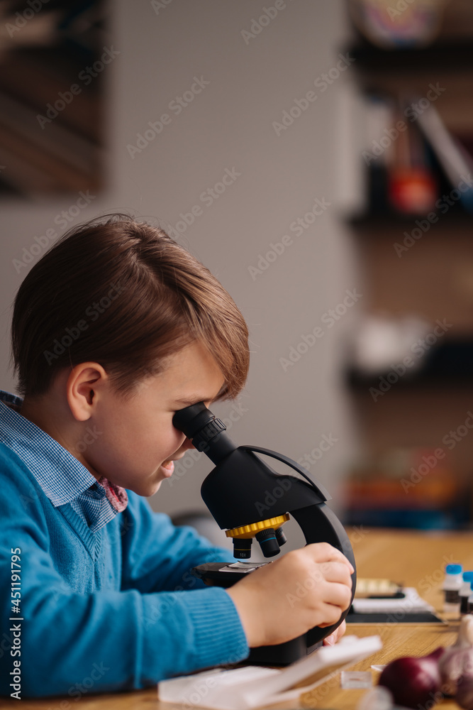 Distance online education, internet learning. First grade boy studying at home using microscope, making notes, biology online lesson