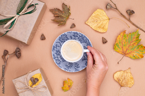 Autumn composition. A female hand holds a cup of coffee, gifts in craft paper, autumn leaves on a beige background. Flat lay, top view