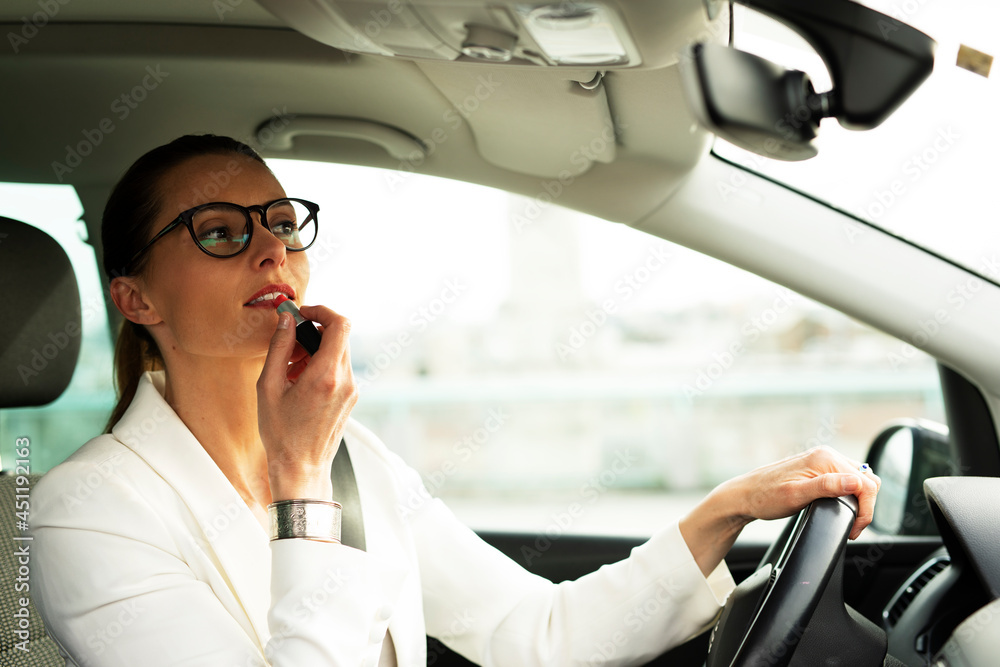 Beautiful young businesswoman sitting behind a wheel of a car doing make up. Attractive businesswoman preparing for the meeting.