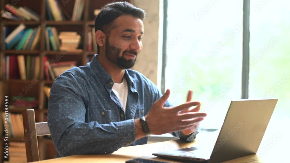 Positive smiling Indian freelancer guy using laptop for virtual meeting at office or library, holding virtual meeting, multiracial eastern male employee waving and greeting online interlocutors