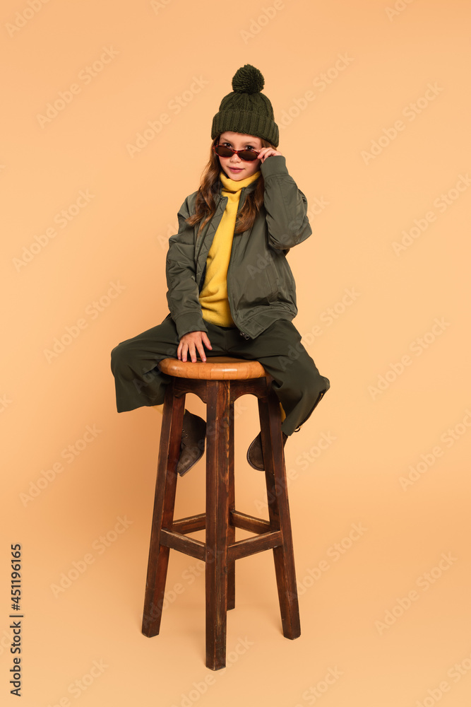 Fototapeta premium trendy kid looking at camera over sunglasses while sitting on high wooden stool on beige
