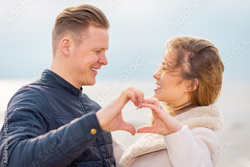Two young couple is making heart, looking at each other and smiling while standing at the beach.