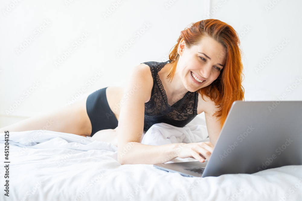 attractive smiling ginger woman middle age lying in bed using laptop in bright room