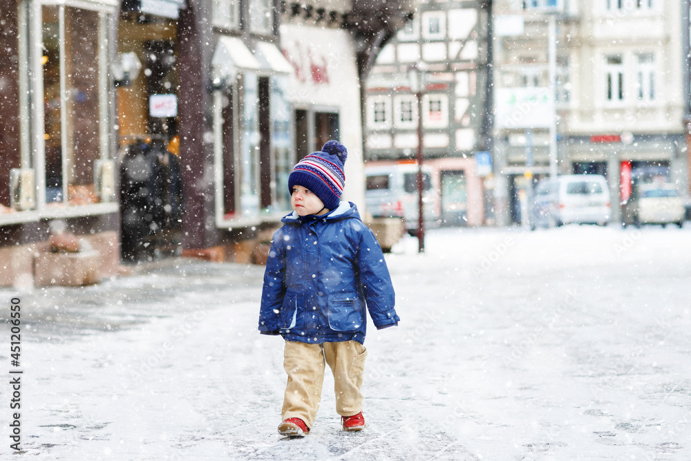 Little toddler boy walking through the snowy city during snowfall. Cute happy child in winter clothes having fun outdoors, on cold day with snow on the street. Little toddler boy walking through the snowy city during snowfall. Cute happy child in winter clothes having fun outdoors, on cold day with snow on the street.