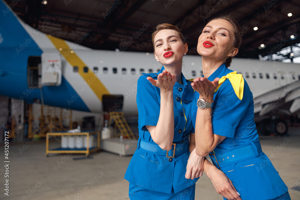 Two air stewardesses in stylish blue uniform blowing kisses to camera, standing together in front of passenger aircraft in hangar at the airport. Occupation concept
