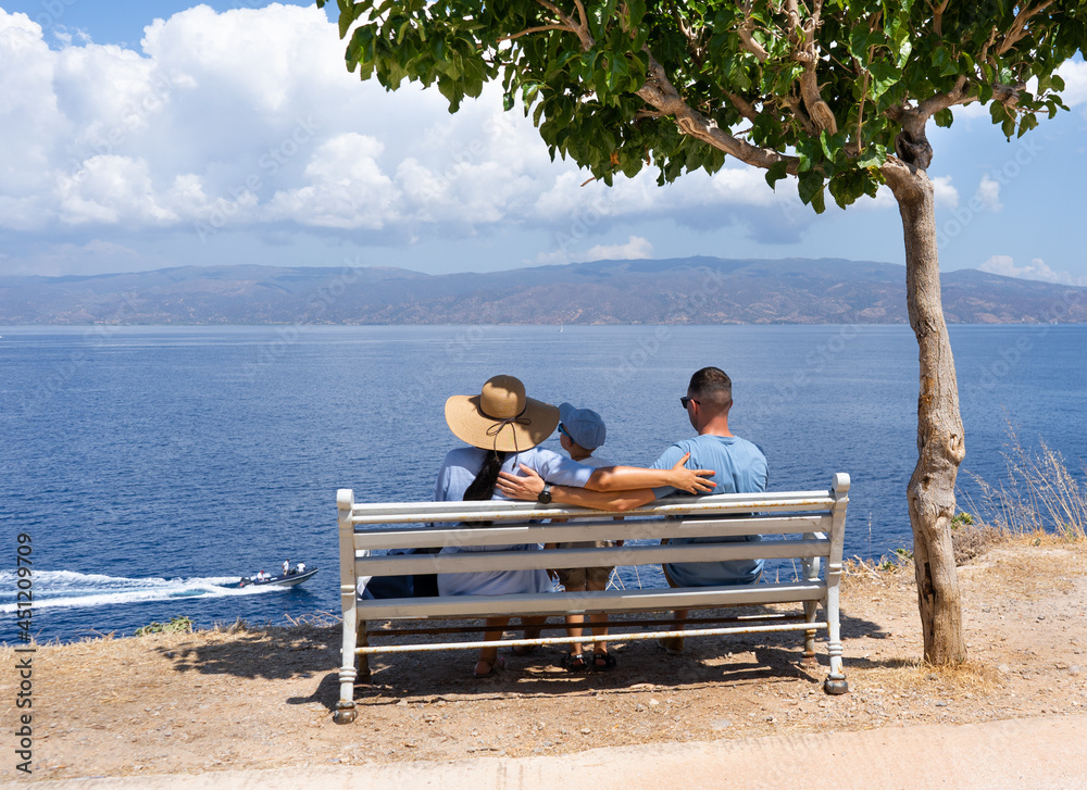 Happy family sits on a bench in the shade of a tree and looks at the sea. Holidays and vacation at sea concept 
