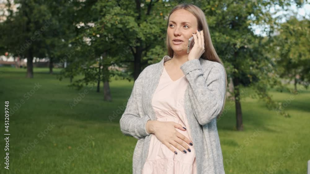 Joyful pregnant woman is speaking on mobile phone standing in city park on summer day smiling enjoying communication. People and technology concept. Joyful pregnant woman is speaking on mobile phone standing in city park on summer day smiling enjoying communication. People and technology concept.