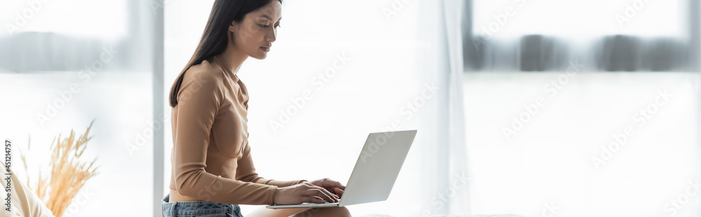 Naklejka premium pretty asian freelancer typing on laptop near window at home, banner.