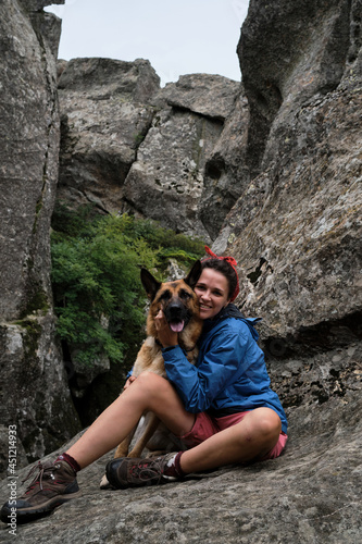 Female tourist on mountain hike in blue jacket and boots. Pretty young Caucasian woman is sitting on rock and hugging dog. Hiking in mountains with German Shepherd.