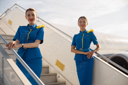 Two beautiful air stewardesses in blue uniform smiling at camera, standing on airstair and welcoming passengers. Aircrew, occupation concept photo