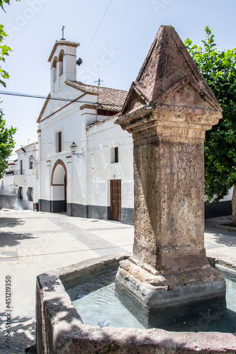 stone fountain with hexagonal pylon and square central column in Yegen de la Alpujarra located in the town square next to the church with bell tower photo