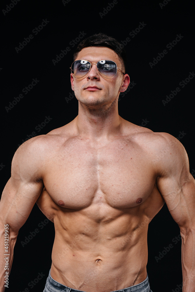 Fototapeta premium A young sexy athlete with perfect abs poses in the studio topless in jeans in a pair of protective glasses. Healthy lifestyle, proper nutrition, training programs and nutrition for weight loss.