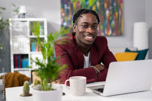 Dark-skinned boy elegantly dressed in jacket sits at desk with a laptop and cup of coffee in the room, smiles at camera showing white teeth, works in a customer service office, financial advisor