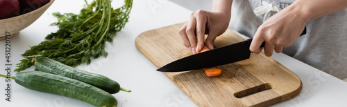 cropped view of woman cutting raw carrot on chopping board near fresh cucumbers on table, banner.