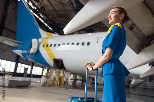 Pretty air stewardesses in bright blue uniform smiling away while standing with suitcase in front of passenger aircraft. Occupation concept