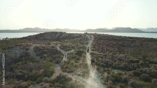 Aerial drone point of view two young adult caucasian women rides on horses outdoor. Embalse de La Pedrera lake view, Orihuela. Spain. Concept of hobby, equestrian sport and active lifestyle photo