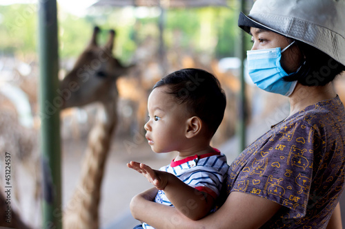 Happy mother wearing mask and Little kid boy watching giraffe in zoo. Happy child having excited with animals safari park on warm summer day. New Normal 