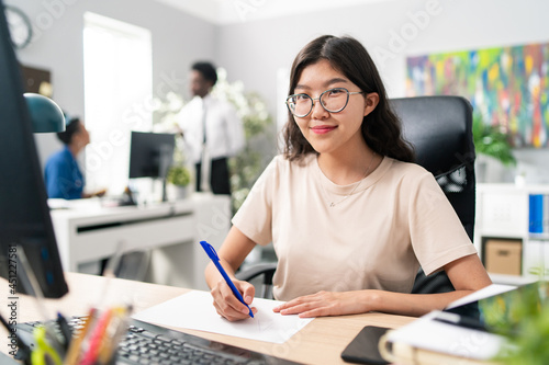 Smiling girl of Asian Korean beauty dressed in loose T-shirt and glasses sits at desk in front of computer working on new project, she holds piece of paper and pen in front of her drawing patterns