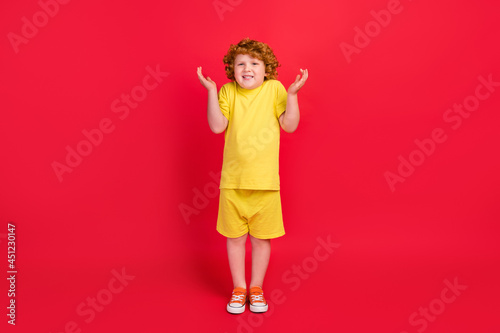 Photo of excited clueless ginger little boy shrug shoulders wear yellow t-shirt isolated red color background