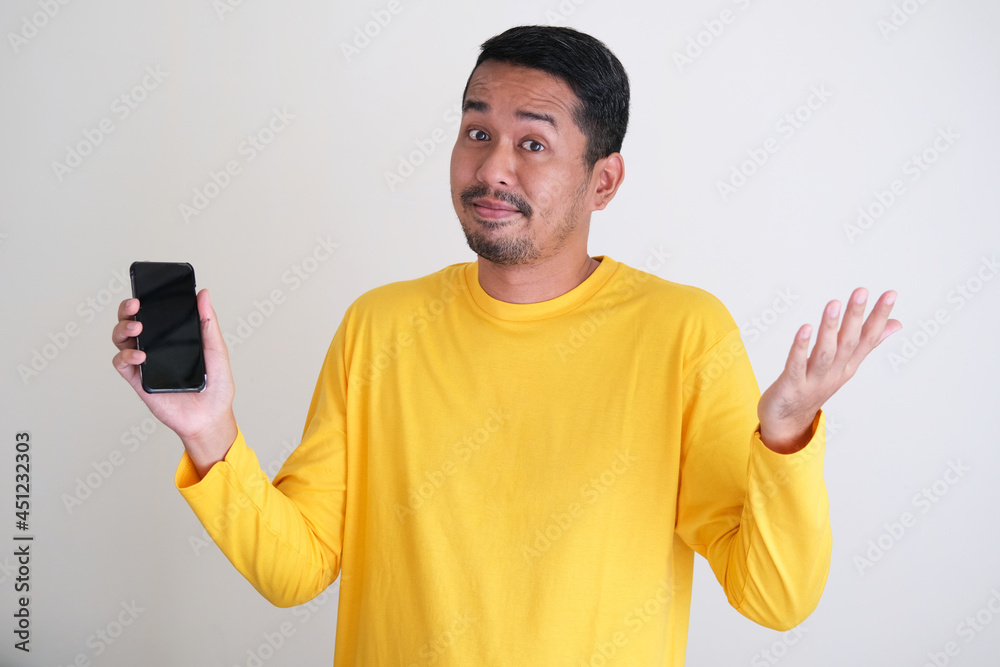 Adult asian man wearing bright yellow clothes holding mobile phone and showing confused gesture Adult asian man wearing bright yellow clothes holding mobile phone and showing confused gesture