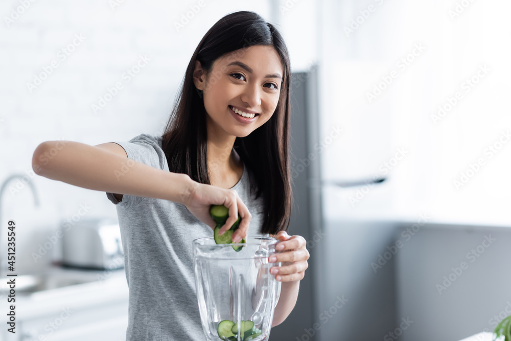 young asian woman smiling at camera while adding sliced cucumber into blender. young asian woman smiling at camera while adding sliced cucumber into blender.