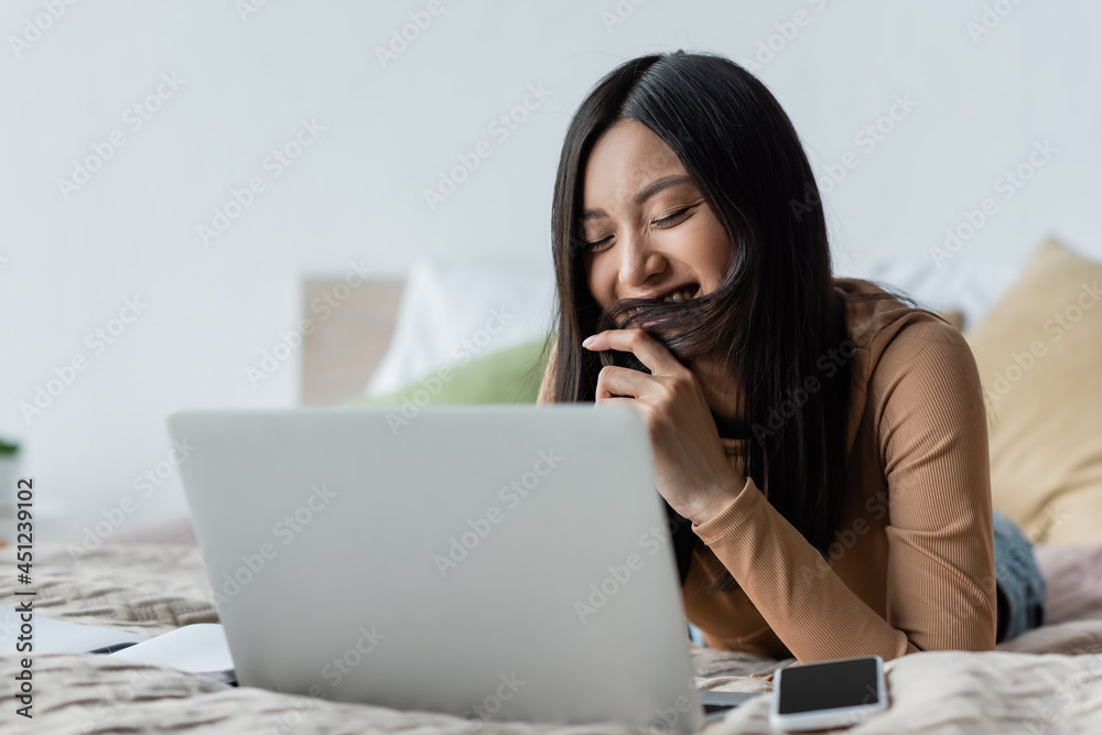 smiling asian woman covering mouth with hair near notebook in bedroom. smiling asian woman covering mouth with hair near notebook in bedroom.