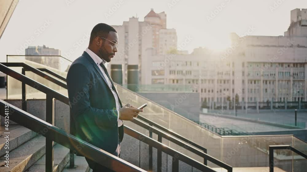 Confident african american businessman leaving office building in evening, networking on cellphone, moving downstairs