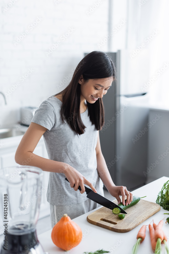 happy asian woman cutting cucumber near fresh carrots and pumpkin in kitchen. happy asian woman cutting cucumber near fresh carrots and pumpkin in kitchen.