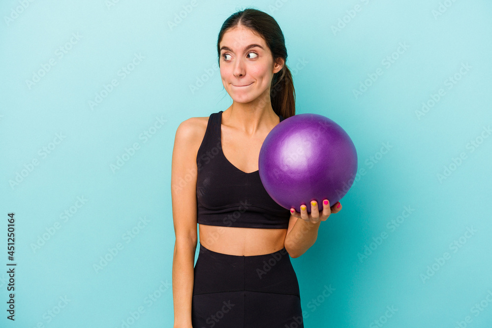 Young caucasian fitness woman holding a ball isolated on blue background confused, feels doubtful and unsure. Young caucasian fitness woman holding a ball isolated on blue background confused, feels doubtful and unsure.