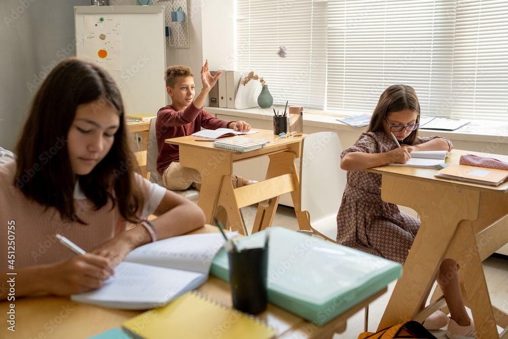 Clever schoolboy raising hand among his classmates making notes in copybooks