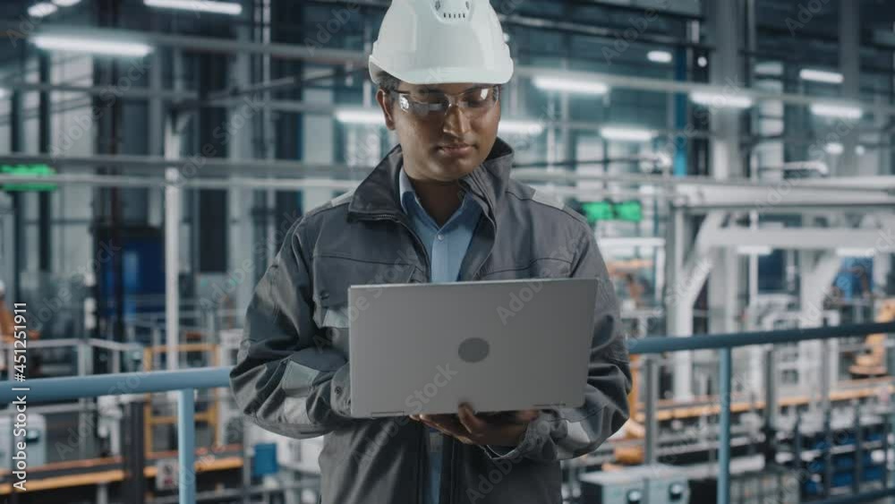 Portrait of Automotive Industry Engineer in Safety Uniform Using Laptop at Car Factory Facility. Happy Assembly Plant Multiethnic Man Specialist Working on Manufacturing Modern Electric Vehicles. Portrait of Automotive Industry Engineer in Safety Uniform Using Laptop at Car Factory Facility. Happy Assembly Plant Multiethnic Man Specialist Working on Manufacturing Modern Electric Vehicles.