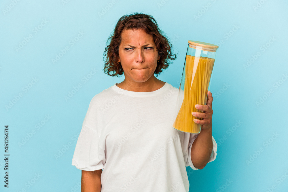 Middle age latin woman holding pasta jar isolated on blue background confused, feels doubtful and unsure.
