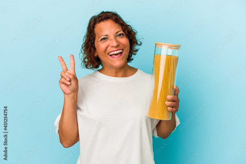 Middle age latin woman holding pasta jar isolated on blue background joyful and carefree showing a peace symbol with fingers. Middle age latin woman holding pasta jar isolated on blue background joyful and carefree showing a peace symbol with fingers.